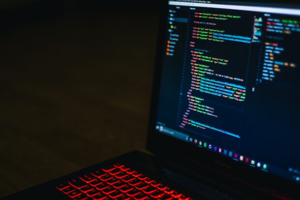 A laptop resting on a table with the screen open showing lines of computer coding. The background is dark and the laptop's keyboard is backlit in red. 