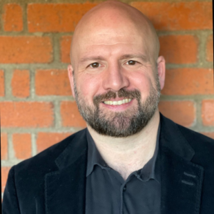 Charles Baird smiling at the camera with a brick wall behind him