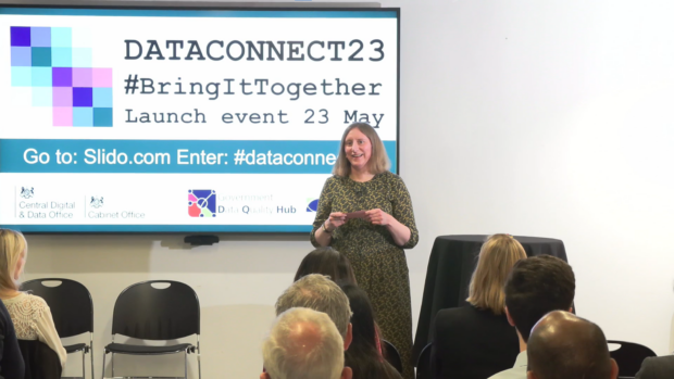Sue Bateman talking to group of people with a sign about DataConnect23 behind her. The sign says 'DataConnect23, #BringItTogether, Launch event 23 May, go to Slido.com and provides the password to enter which is being blocked by Sue in the image.